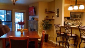 A dining area with a wooden table and chairs, a chandelier, and a kitchen with wooden cabinets and a microwave.