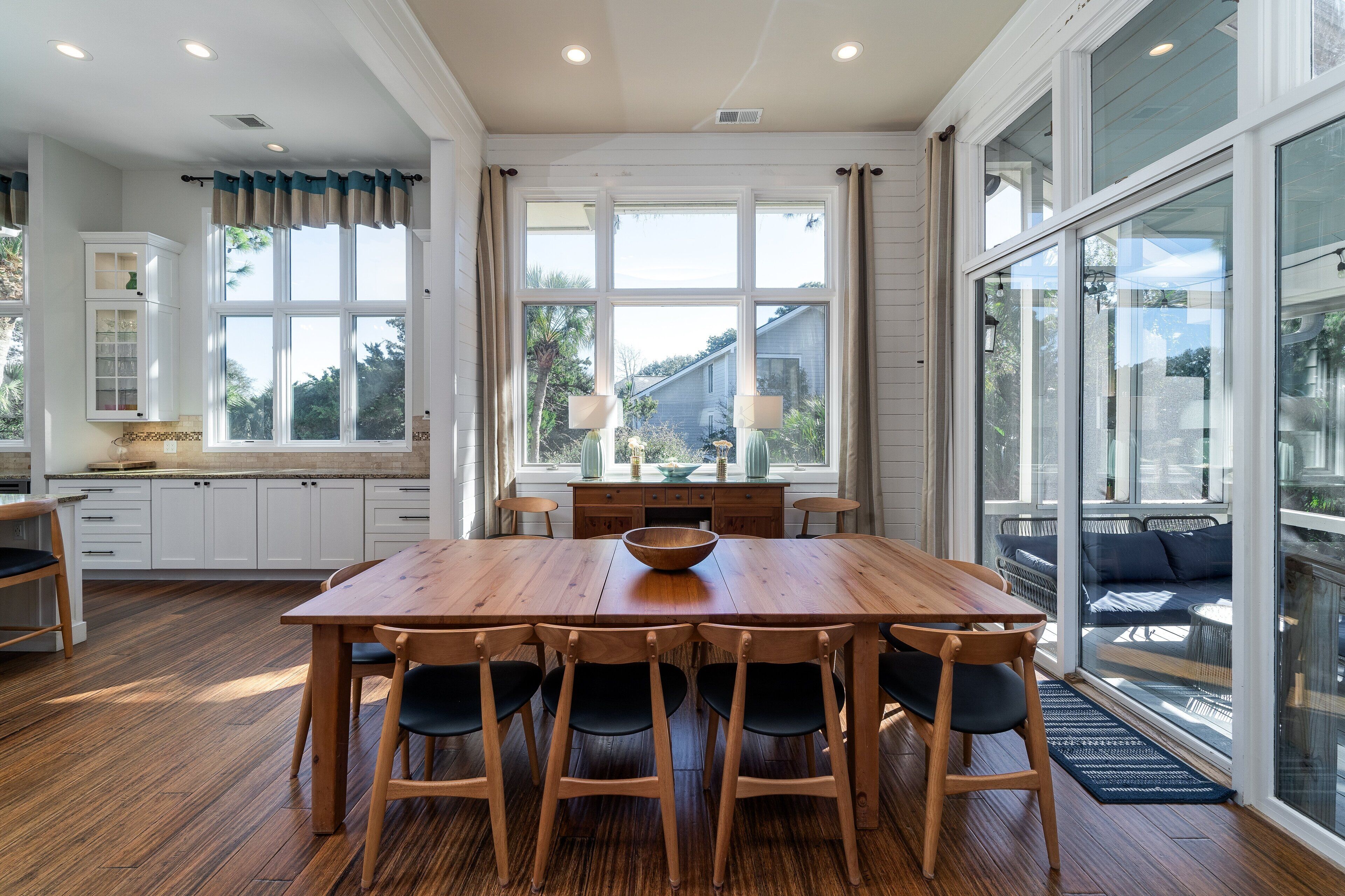 A spacious dining area with a wooden table and chairs, large windows with curtains, and a view of the outdoors.
