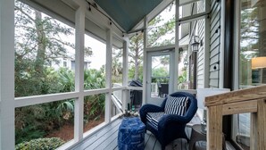 Une véranda couverte avec une terrasse en bois, un pouf bleu à motifs, une chaise à rayures et une vue sur la verdure environnante.