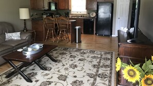 A living room with a sofa, coffee table, and a rug. There is a kitchen area with wooden cabinets, a refrigerator, and a dining table with chairs. Sunflowers are on a table in the foreground. - Beautiful rural setting cottage (Galax)
