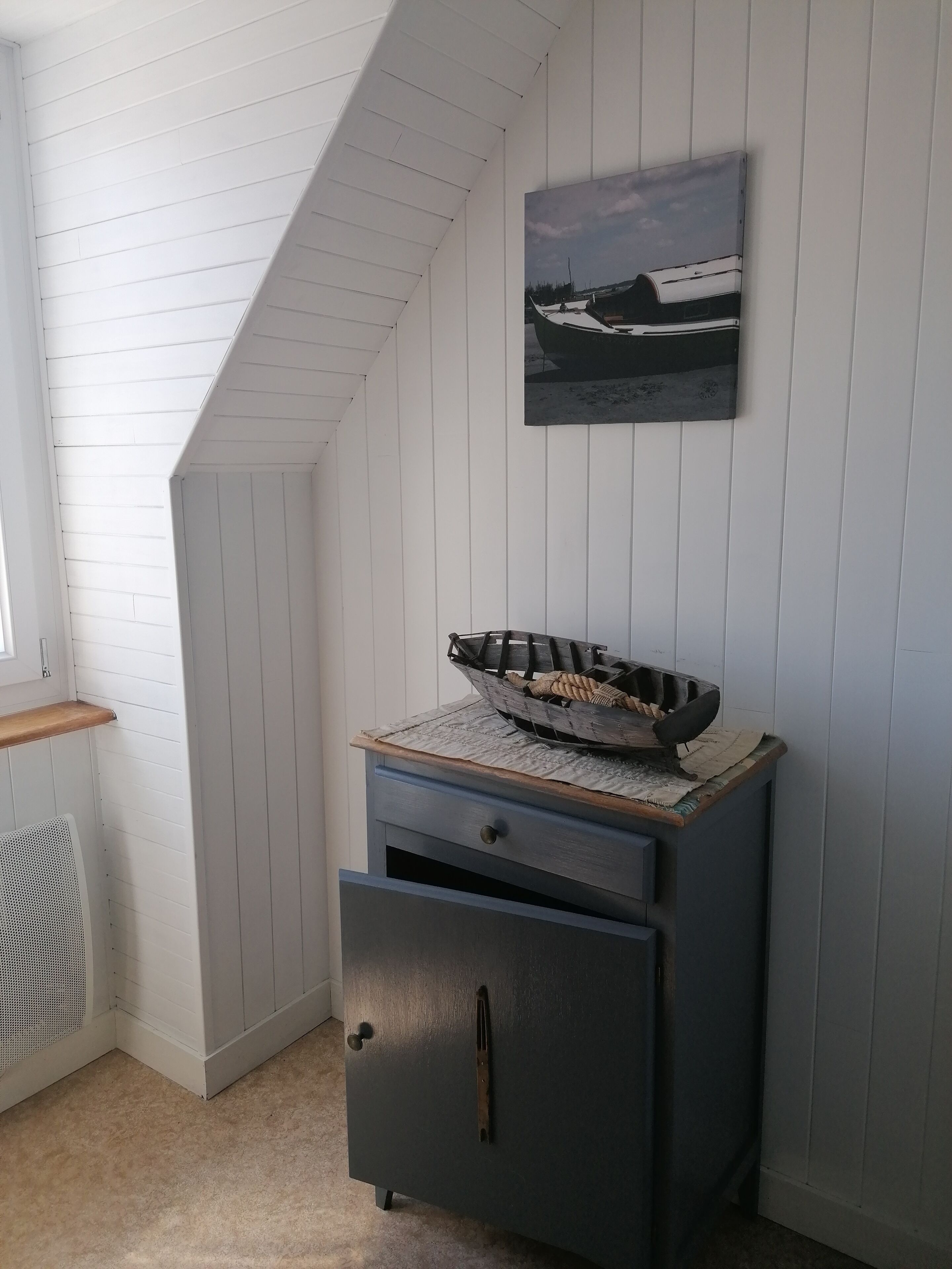 Ett hörnrum med ett sluttande tak, en liten hylla i trä, ett mörkt skåp med en låda och en inramad tavla på väggen.