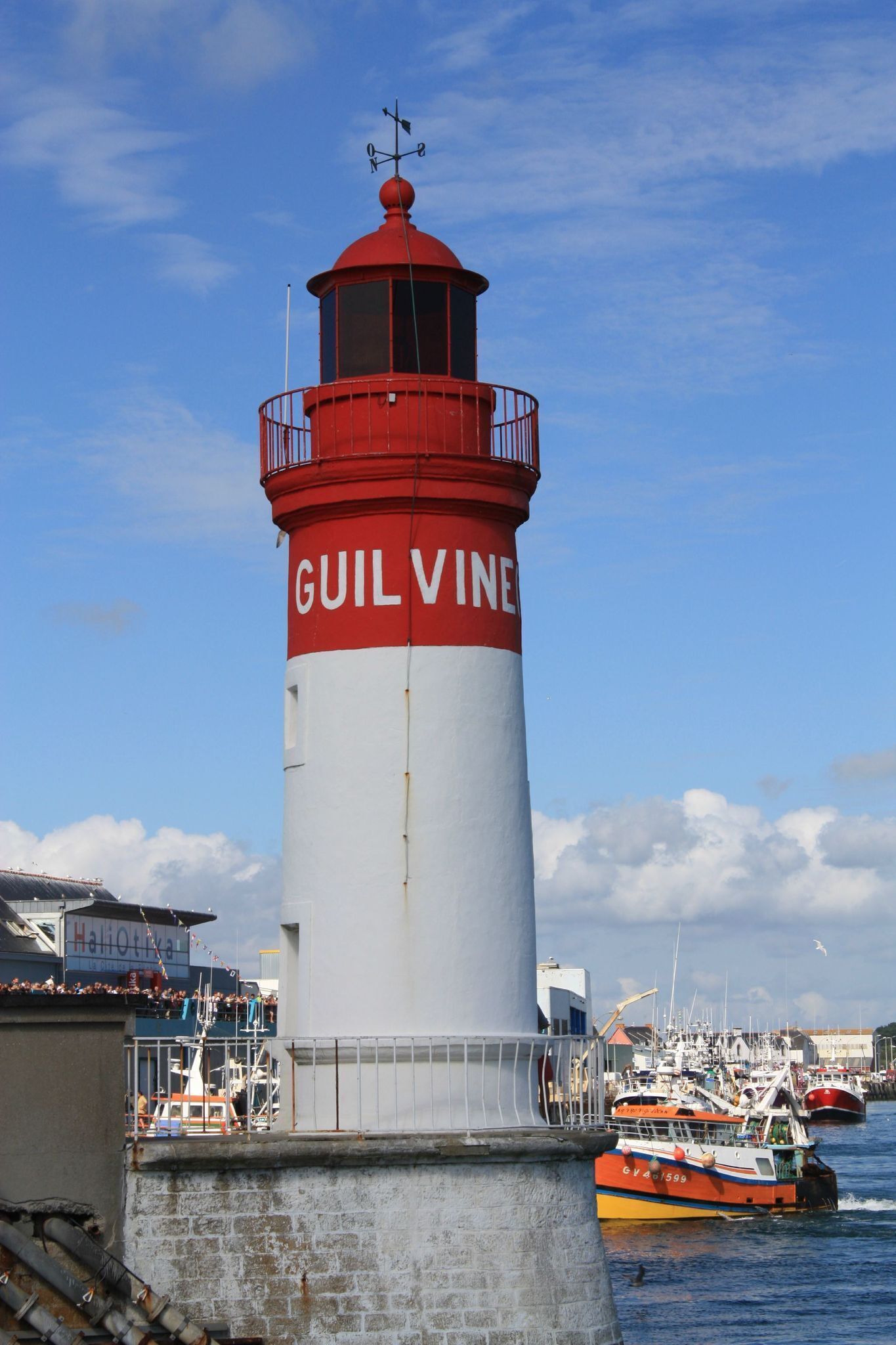 Een vuurtoren genaamd "GUILVINE" met een rood-wit kleurenschema, gelegen nabij een haven met boten en gebouwen op de achtergrond.