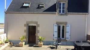 A building with a dark roof, a central door, and two windows with shutters. There are potted plants and outdoor furniture. - Fisherman's house a few meters from the beach (Guilvinec)