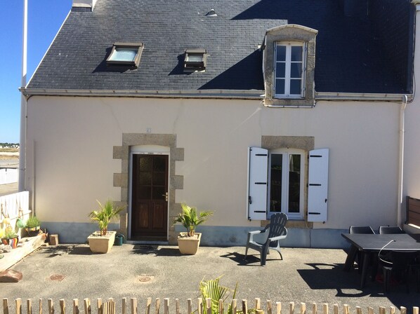 A building with a dark roof, a central door, and two windows with shutters. There are potted plants and outdoor furniture.