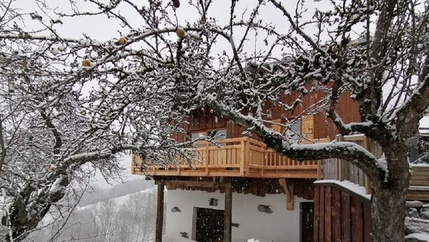 A wooden chalet with a balcony, surrounded by snow-covered trees and a snowy landscape.