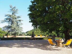 Un espace extérieur aménagé avec des chaises jaunes et une table en pierre, entouré d’arbres et d’un mur de pierre.