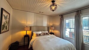 Une chambre à coucher avec un lit, deux lampes de chevet, une estampe botanique encadrée et une fenêtre avec des rideaux.