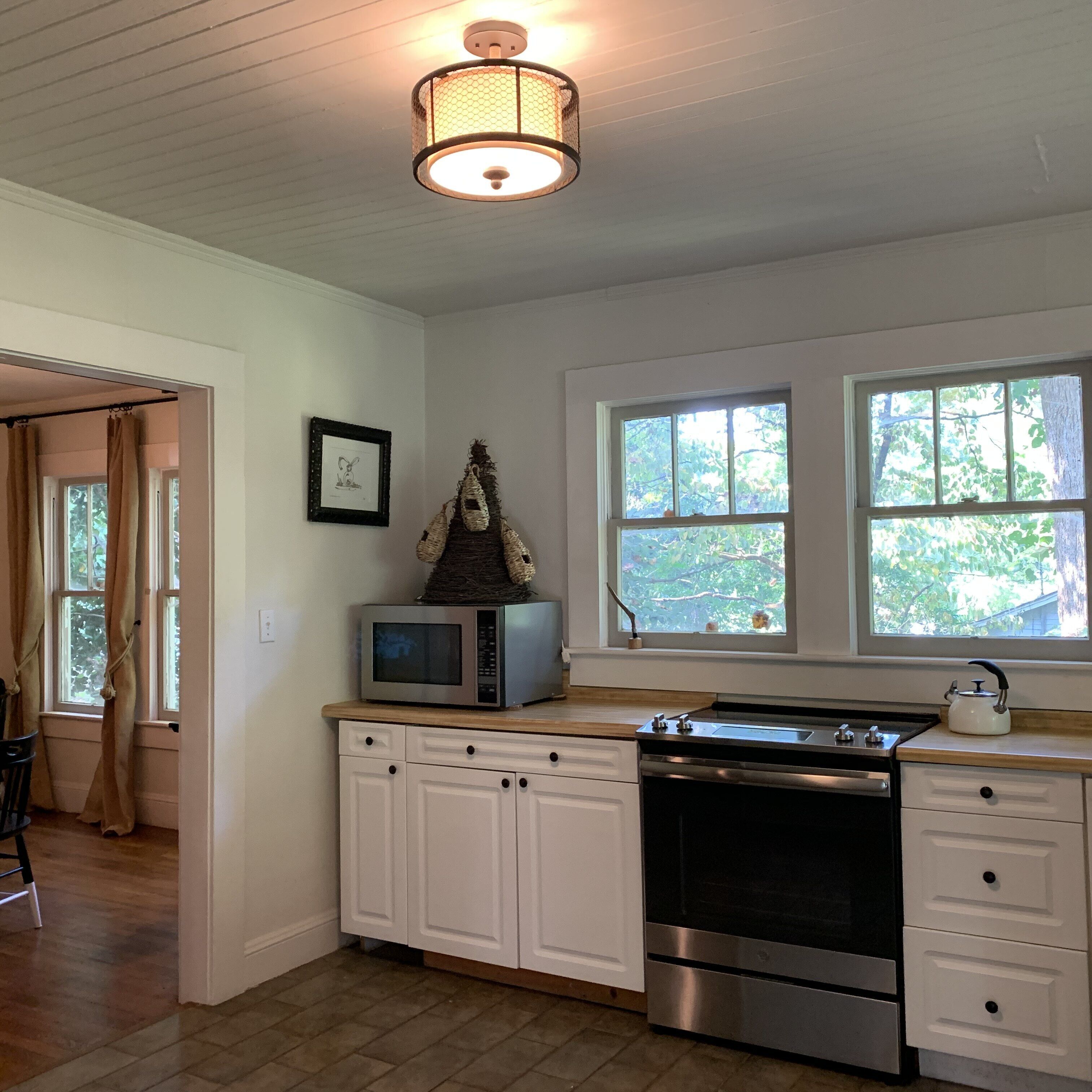 A kitchen with white cabinets, a microwave, a stove, and a hanging light fixture. There are windows with views of trees and a door leading to another room.
