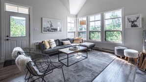 Ein modernes Wohnzimmer mit einem grauen Sofa, einem Couchtisch und einem Hund. Große Fenster und ein Deckenventilator sind vorhanden.