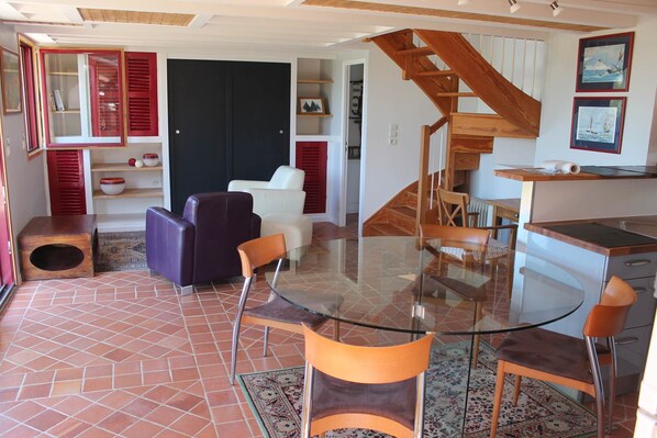 Una sala moderna con una mesa de comedor de vidrio, dos sofás morados, una escalera de madera y una zona de cocina con encimera de madera.