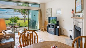 Ein geräumiges Wohnzimmer mit einem großen Schiebeglasfenster, das auf eine Terrasse führt, einem Kamin, einem Fernseher und einem Essbereich mit Tisch und Stühlen.