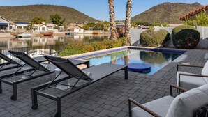 A pool area with lounge chairs, a hot tub, and a view of a lake and hills.