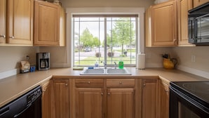 A kitchen with wooden cabinets, a black dishwasher, a coffee maker, and a window with a view of a street and trees.