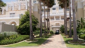 Een resort met een centrale pad, omzoomd door palmbomen en een verzorgde tuin, dat leidt naar een gebouw met meerdere balkons en een overdekte gang.