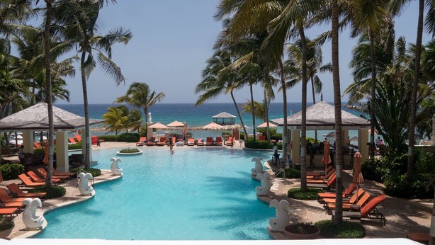 Een zwembadgedeelte van een resort met ligbedden, palmbomen en prieel.