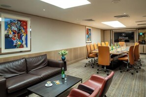 A conference room with a brown leather sofa, a coffee table, and a floral vase. There are multiple chairs around a table, and a large screen displaying the Hilton logo. - The Vista at Hilton Tel Aviv (Tel Aviv)