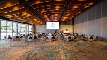 Un salón de conferencias con mesas y sillas redondas, una pantalla grande mostrando el logotipo de Hilton y una alfombra con un diseño circular.