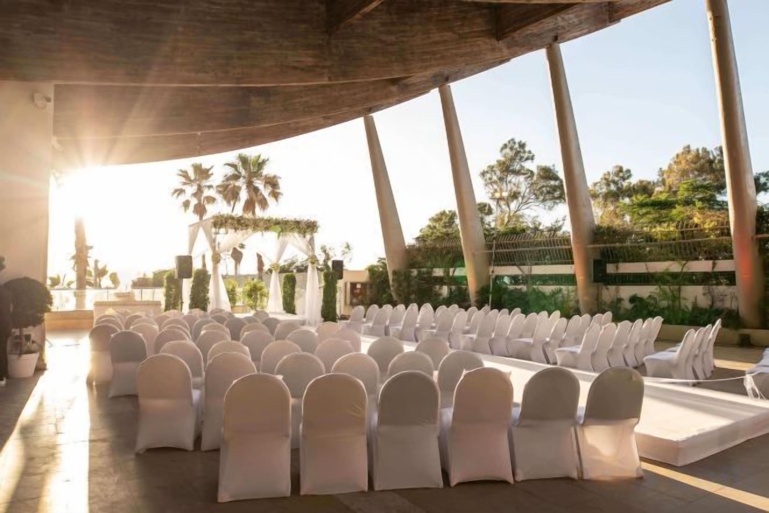 An outdoor seating area with rows of chairs and a stage, surrounded by palm trees and a clear sky.