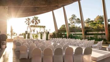 An outdoor seating area with rows of chairs and a stage, surrounded by palm trees and a clear sky.