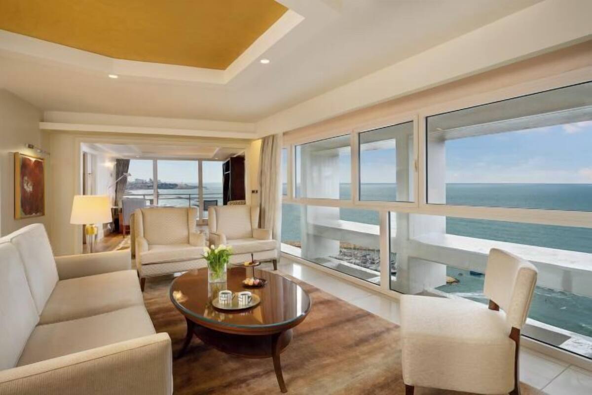 Een hotelkamer met een groot raam met uitzicht op zee, een ronde salontafel en twee banken.