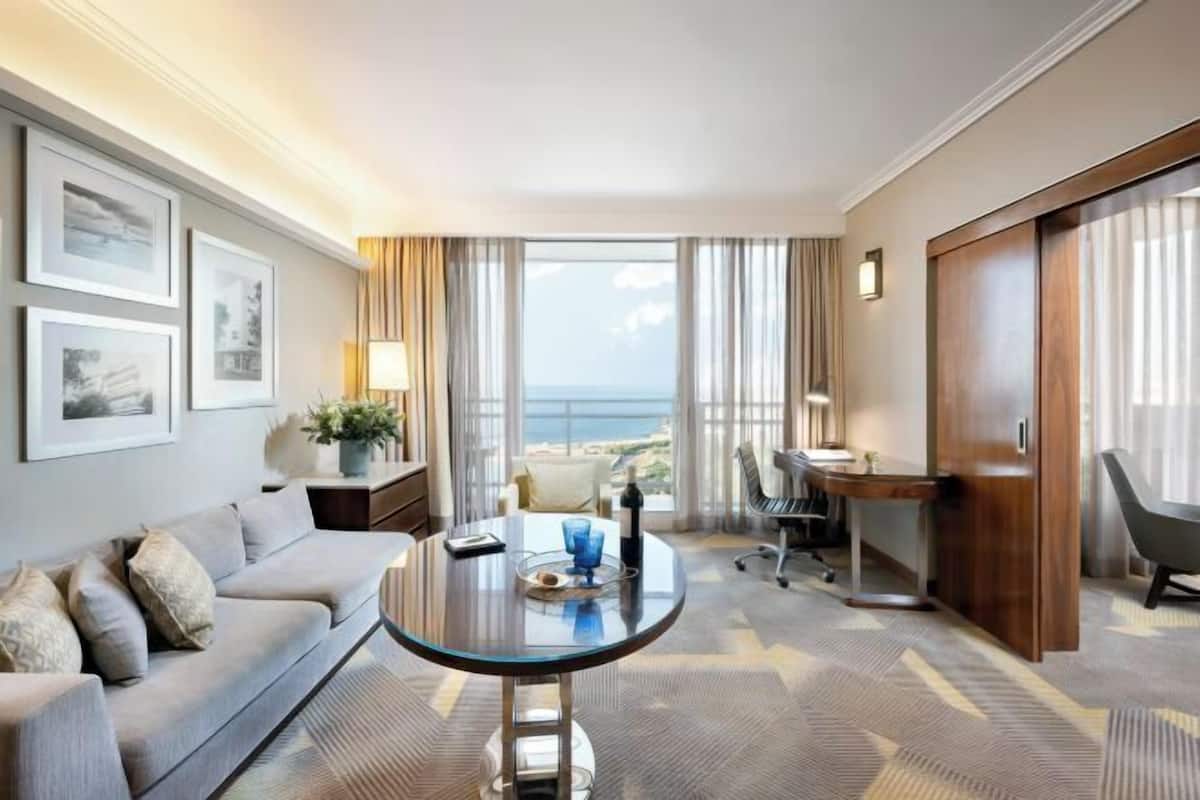 Een hotelkamer met een bank, een glazen tafel, een bureau, een stoel, een lamp, uitzicht op zee en ingelijste foto's aan de muur.