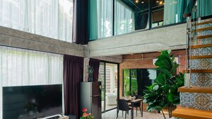 Un vaste espace de vie comprenant un grand téléviseur à écran plat, un canapé gris et une table basse. Un escalier mène à un niveau supérieur, et une grande plante est visible dans un coin.