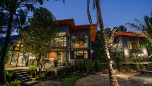Une maison moderne à deux étages, dotée d’un balcon et entourée de plantes tropicales et de palmiers.