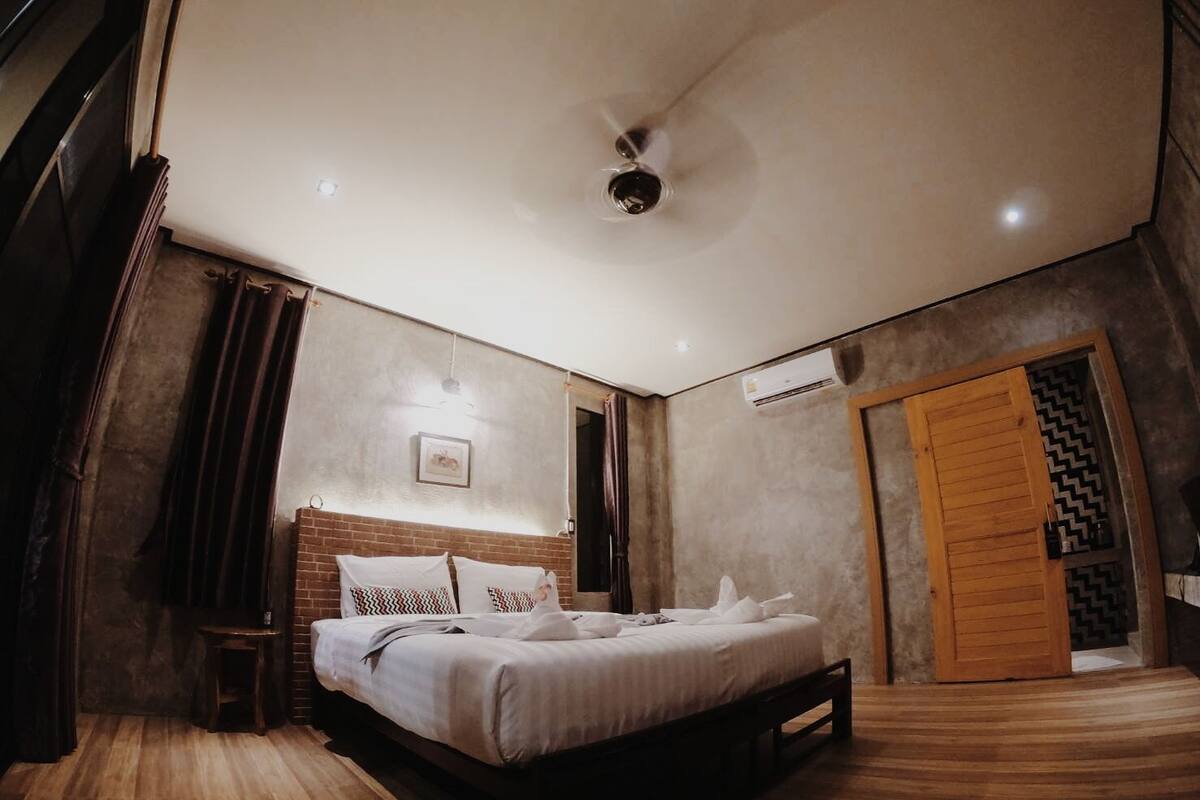 A modern bedroom with a bed, a small stool, a ceiling fan, and a wooden door.