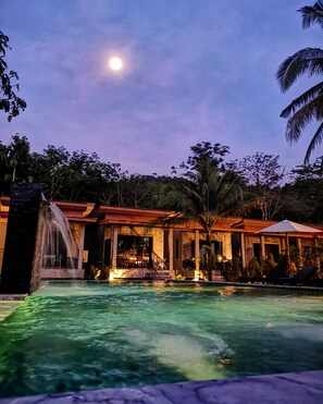 Zona de piscina con cascada, espacio de descanso cubierto y vistas al cielo nocturno.