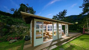 A wooden cabin with a deck, large windows, and a stone wall in the background.