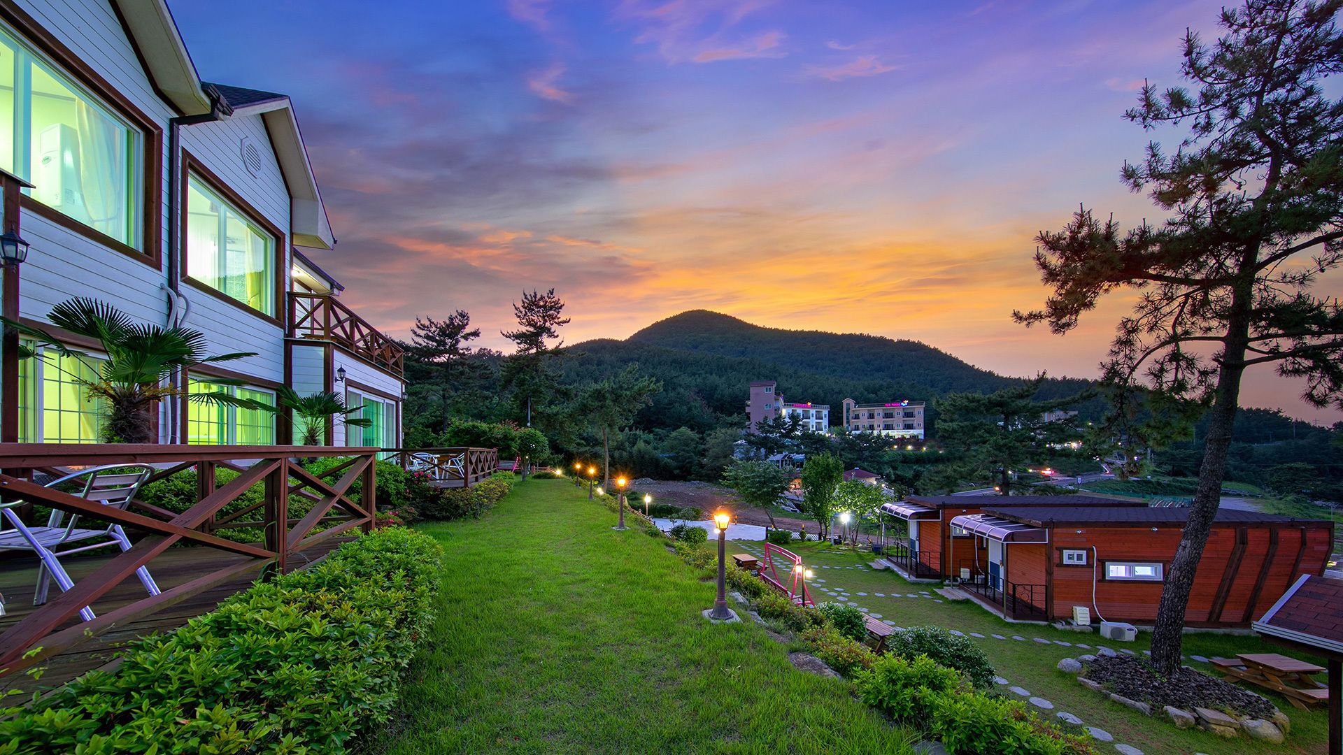A hotel with a well-maintained garden, a deck with chairs, and a view of a mountain at sunset.