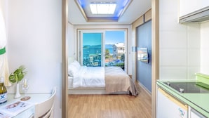 A hotel room with a bed, a desk with a bottle of wine, a plant, and a view of the sea.