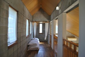 A narrow hallway with a wooden ceiling and stone walls. There are large windows on both sides, and a bed with white linens is visible in the center. - Sea-K (Motobu)