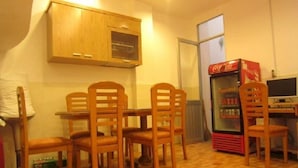 Een klein eetgedeelte met een houten tafel en vier stoelen, een houten kast erboven, een koelkast met Coca-Cola logo, en een raam met uitzicht op buiten.