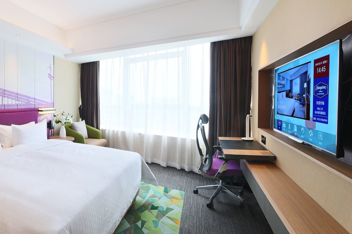 A hotel room with a large bed, a desk, a chair, a TV, and a window with curtains.