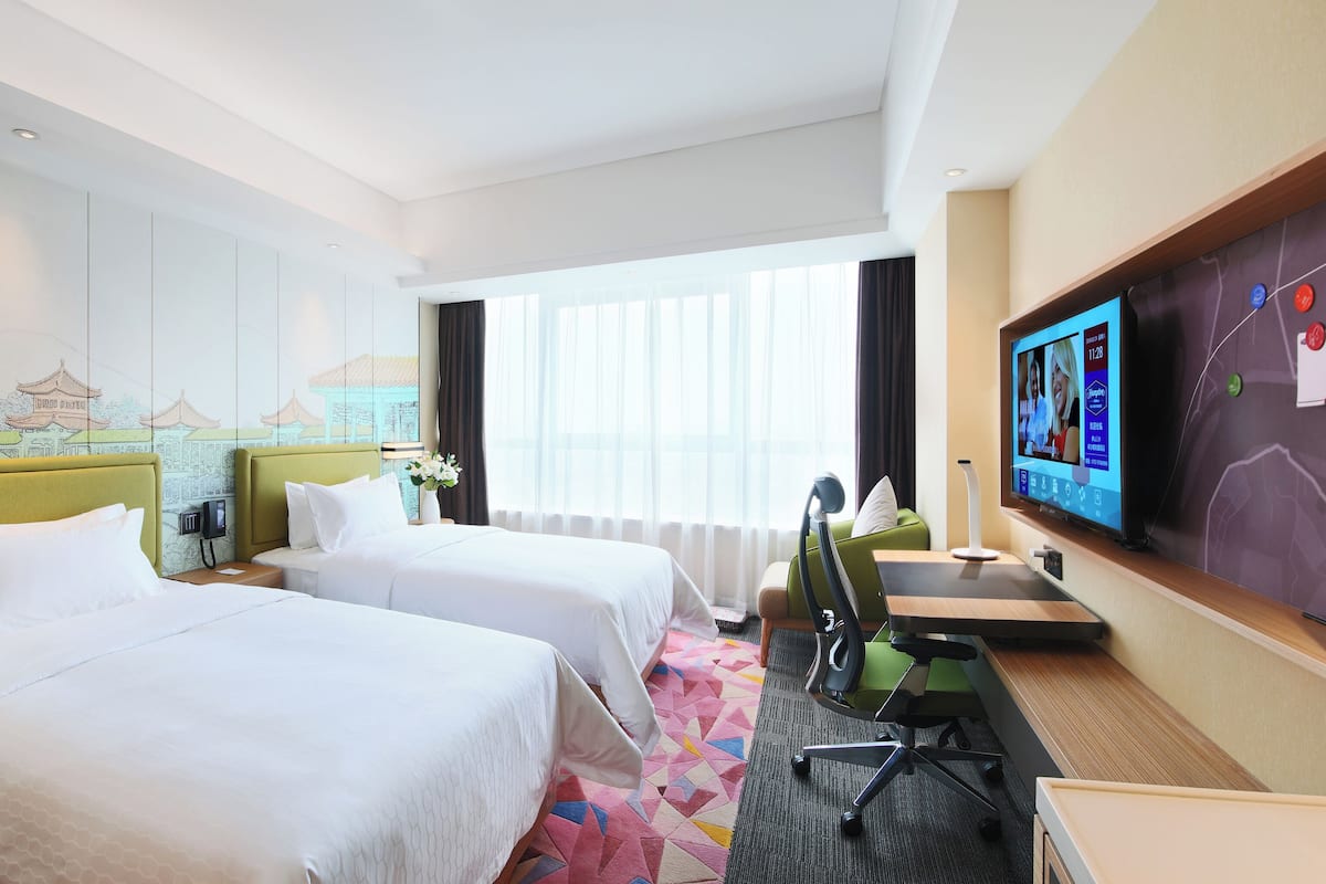 A hotel room with two beds, a desk with a chair, a television, and a large window with curtains.