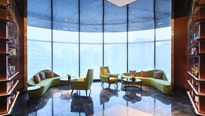 Un amplio salón con un sofá verde curvo, sillones amarillos y una mesa de centro, rodeado de estanterías y grandes ventanales con vistas a la ciudad.