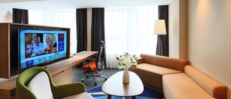 A hotel room with a TV, sofa, chairs, a small table, and a vase of flowers.