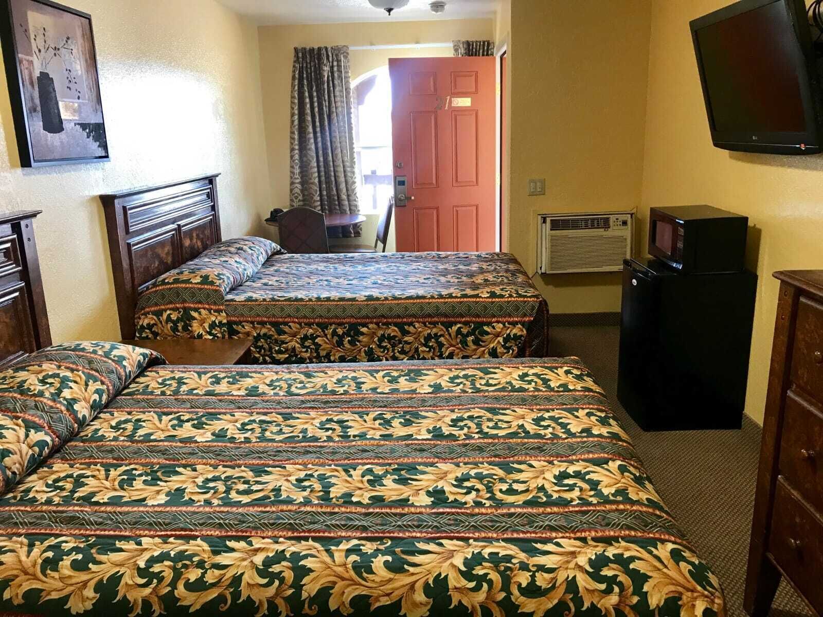 A hotel room with two beds, a television, a refrigerator, and a door leading to a hallway.