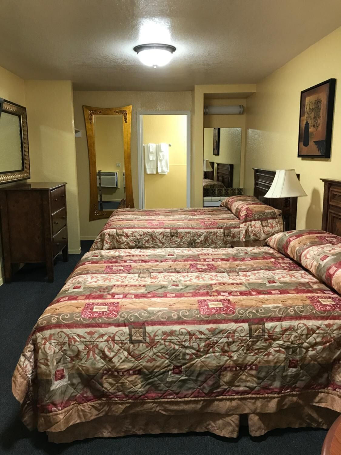 A hotel room with two beds, a dresser, a mirror, a lamp, and a painting on the wall.