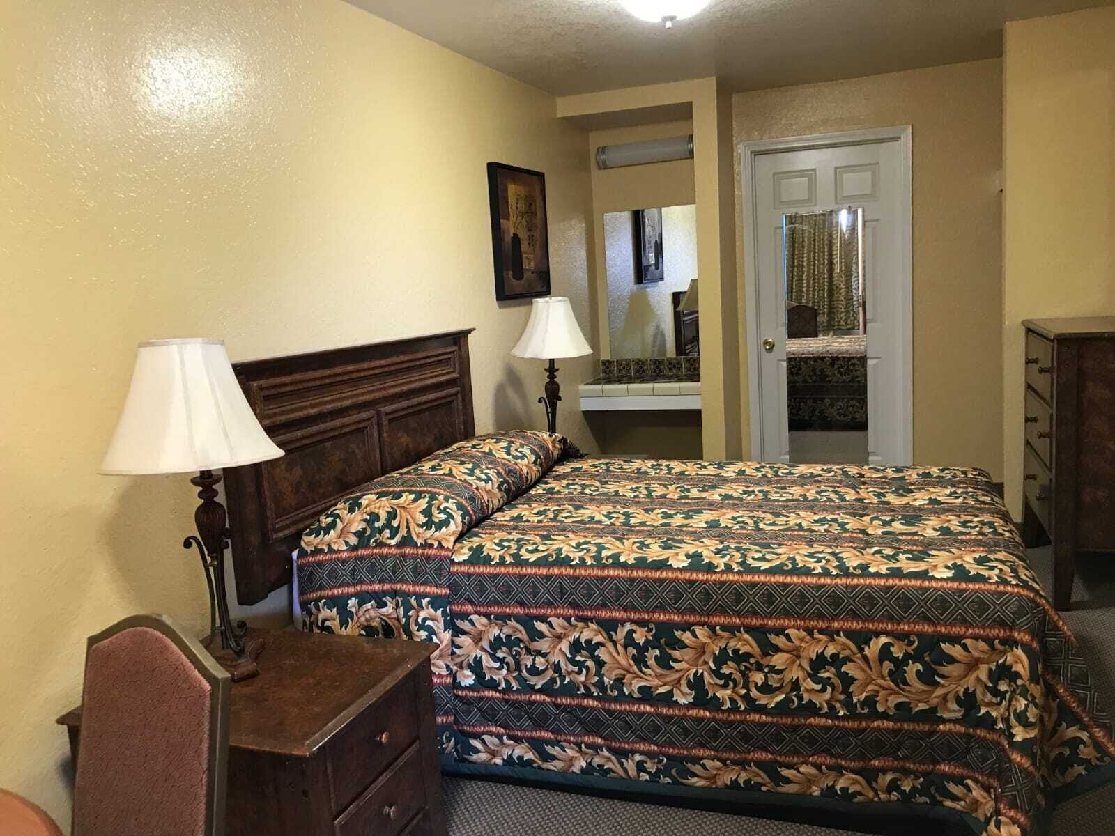 Un dormitorio con una cama, dos lámparas de noche, un tocador, un espejo y una puerta que da a otra habitación.