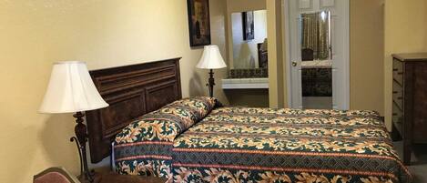 Un dormitorio con una cama, dos lámparas de noche, un tocador, un espejo y una puerta que da a otra habitación.
