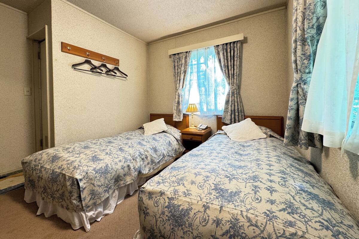 Ein Zimmer mit zwei Betten, einem hölzernen Kopfteil, einem Fenster mit Vorhängen, einem Nachttisch mit Lampe und einem Kleiderständer.