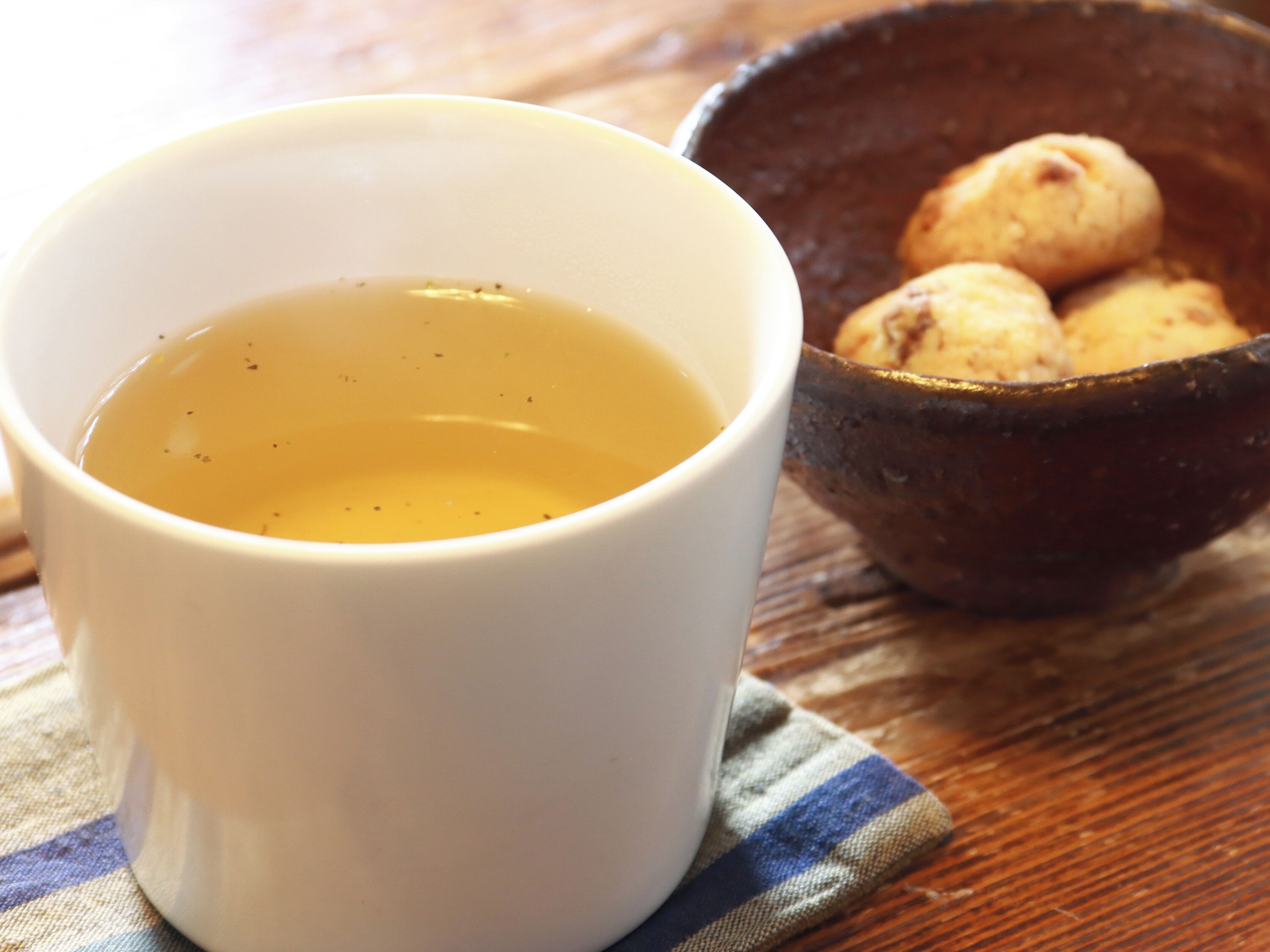 A white cup filled with a yellowish liquid, possibly tea, on a wooden table with a dark bowl containing round, light-colored objects in the background.