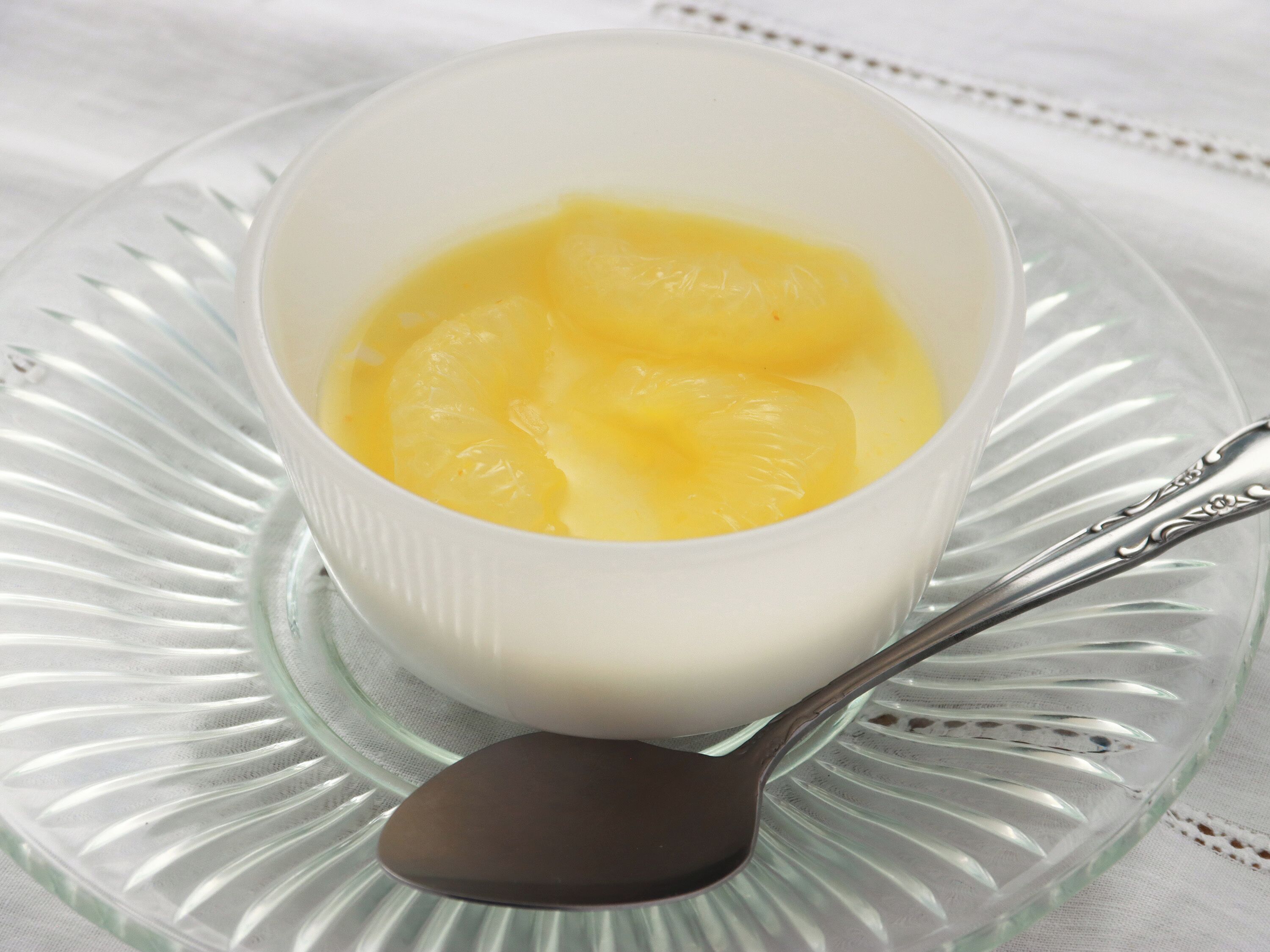 Uma tigela de vidro transparente com interior branco contendo uma substância amarela, possivelmente uma sobremesa, sobre um prato de vidro com uma colher.