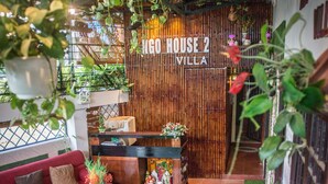 Un espacio al aire libre acogedor con un sofá rojo, una mesa de madera y sillas. Se ve una pared de bambú con la inscripción 'LAGO HOUSE 2 VILLA' al fondo.