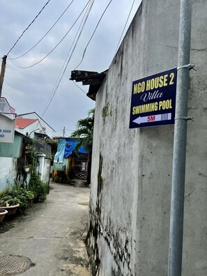 A narrow alley with a concrete wall, a sign for "Ngo House 2 Villa Swimming Pool" pointing to the left, and a blue tarp-covered structure in the background. - Ngo House 2 Villa (Da Nang)