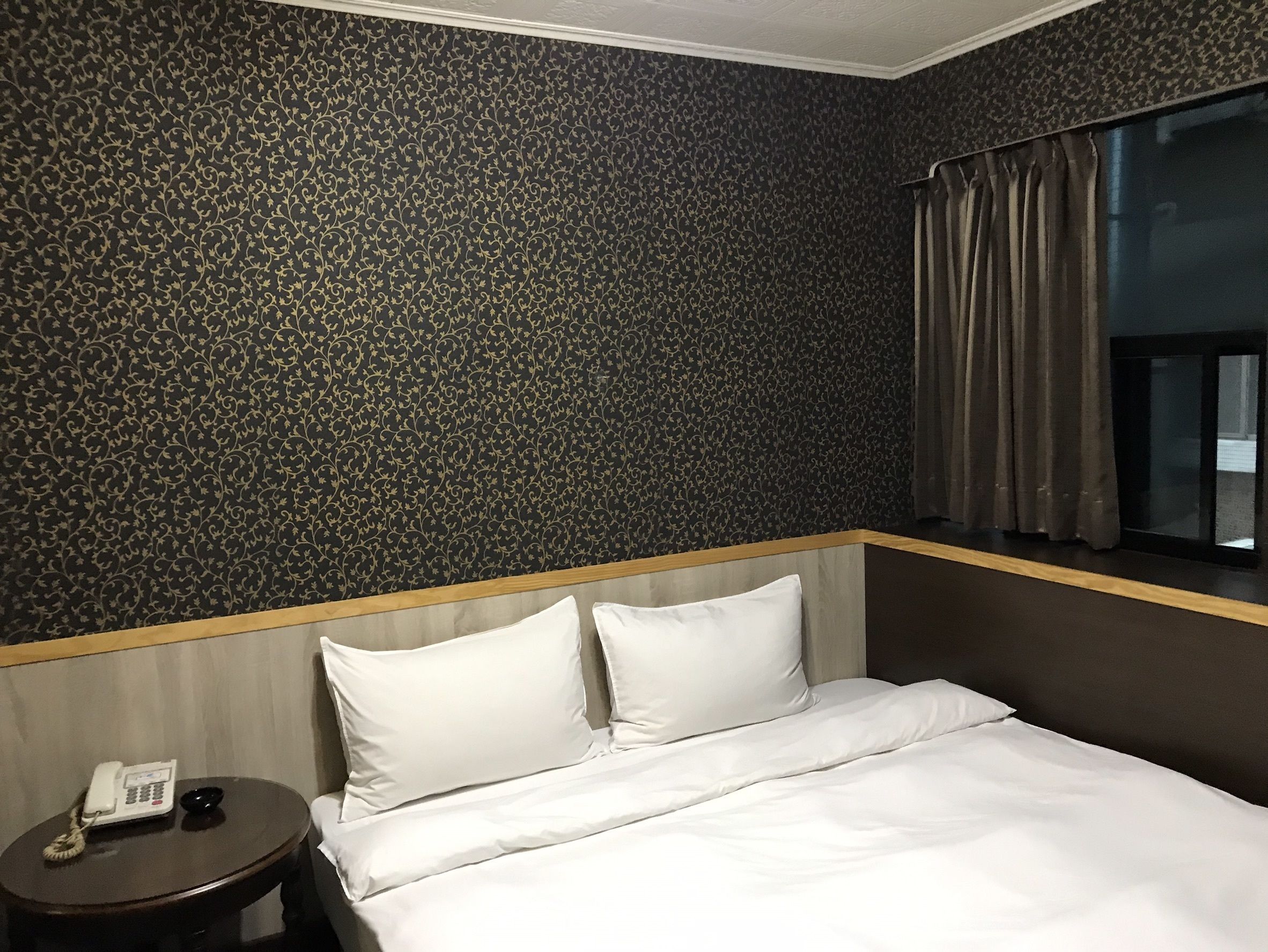 A hotel room with a bed, a bedside table with a phone, and a window with curtains.