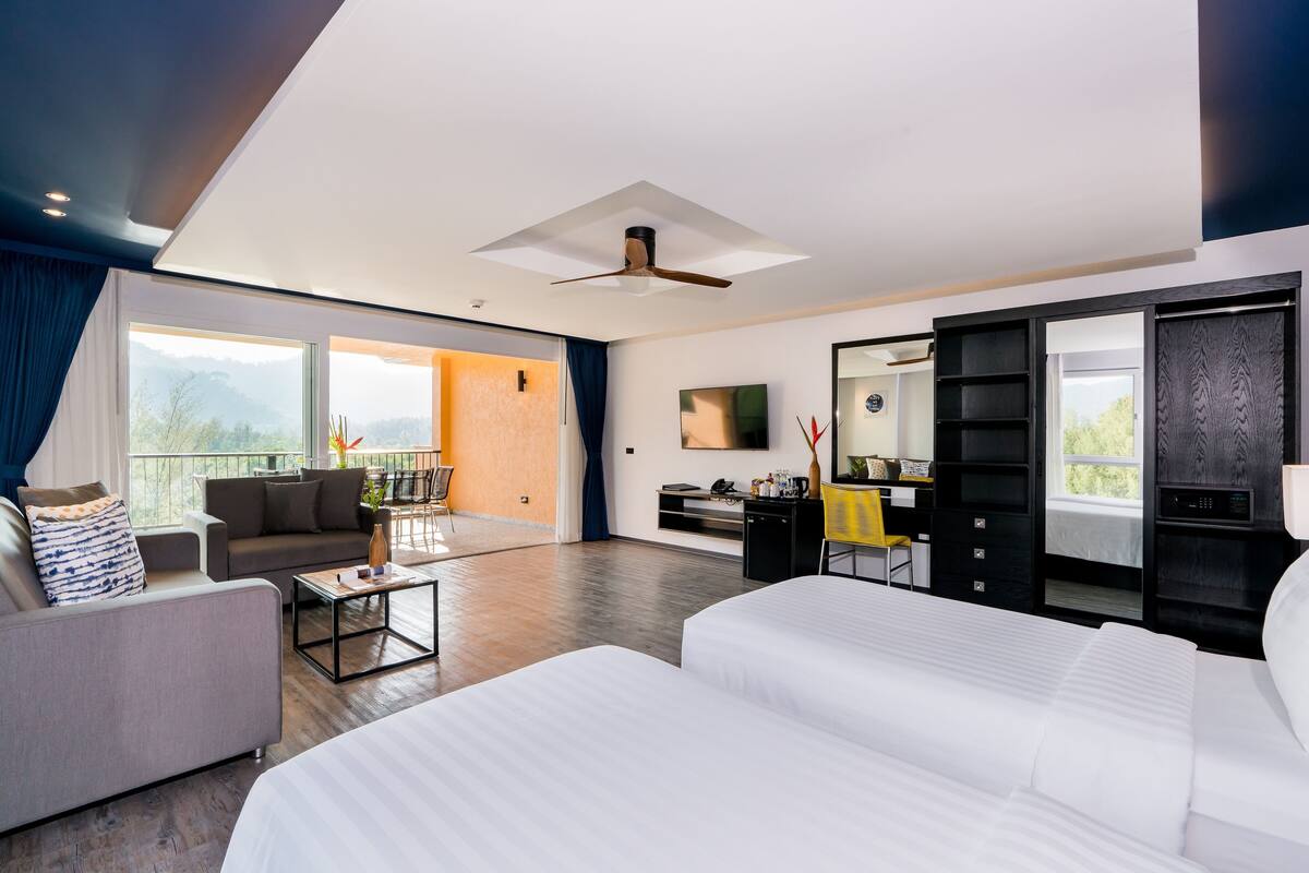 A modern hotel room with a large bed, a sofa, a coffee table, a TV, and a balcony with a view of the mountains.