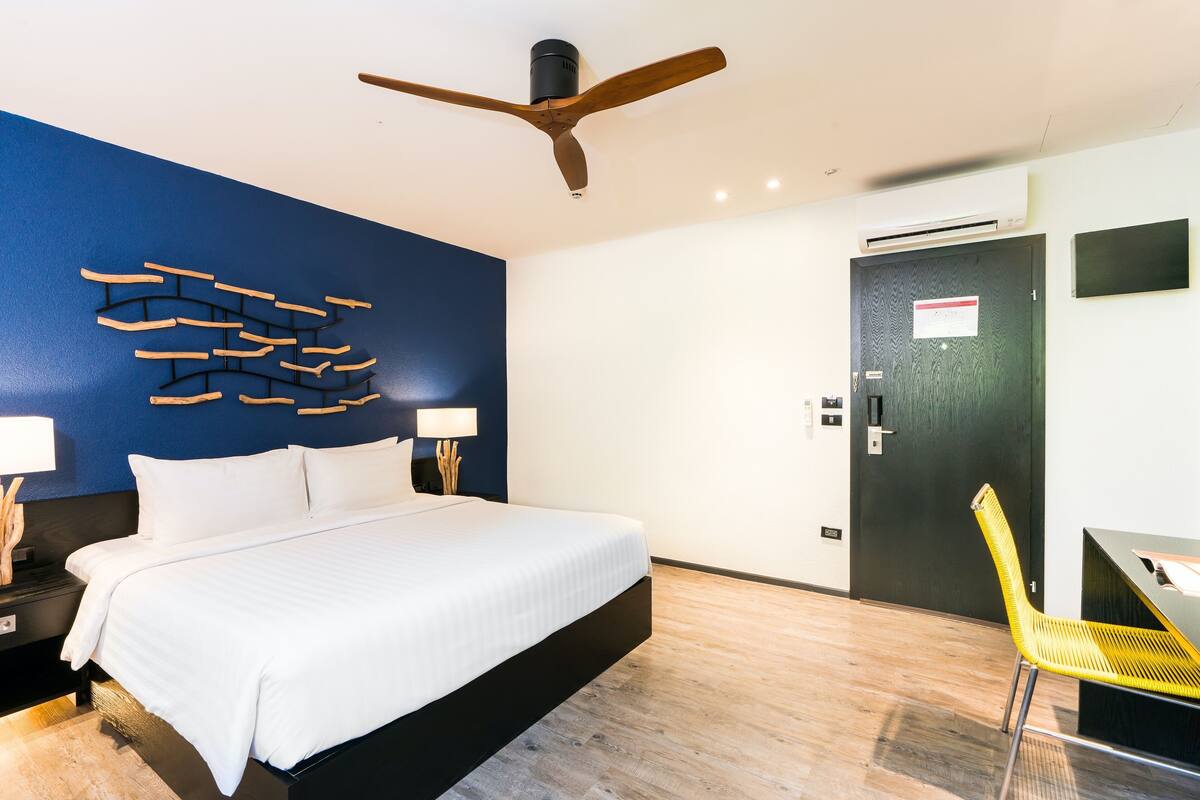 A modern hotel room with a bed, a desk with a chair, a ceiling fan, and a wall-mounted air conditioning unit.